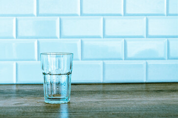 An empty glass water glass stands on a wooden table against a white wall.