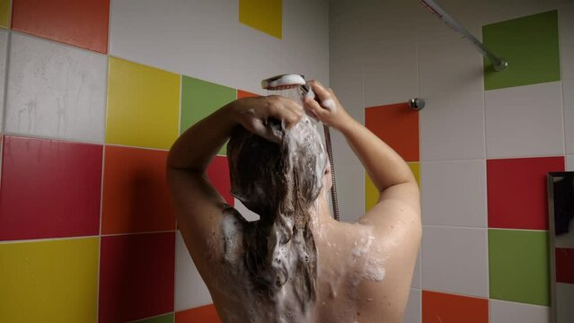 A woman washes her hair. The stream of water from the shower washes the shampoo out of her long, dark hair. Foam flows down her back. Colorful tiles in the background. Hair care concept