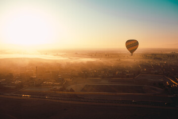 One single hot air balloon in the air, flying low height above the houses. Rearly morning sunrise time, foggy landscape illuminated by sun rays