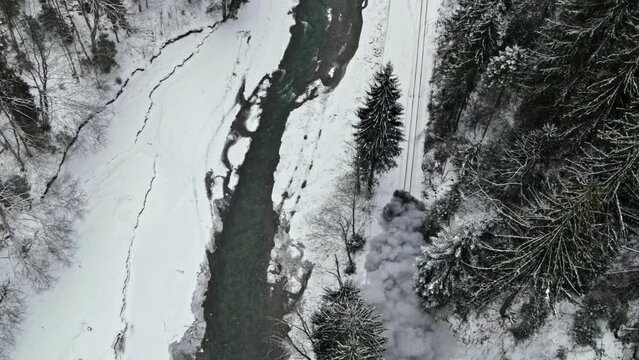 Aerial drone view of the moving steam train Mocanita in a valley along a river in winter, hills covered with bare forest and snow, Romania