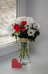 A bouquet of white chrysanthemums with a bush rose, coral in color, stands in a transparent vase with gold rims on the window. In front of the vase is a card in the form of a pink heart.