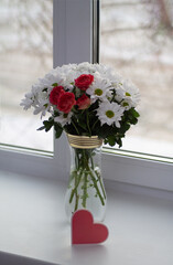 A bouquet of white chrysanthemums with a bush rose, coral in color, stands in a transparent vase with gold rims on the window. In front of the vase is a card in the form of a pink heart. 