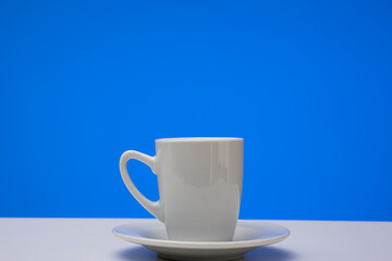 Small white ceramic coffee cup on a plate. Close up studio shot, isolated on blue background