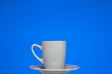 Small white ceramic coffee cup on a plate. Close up studio shot, isolated on blue background