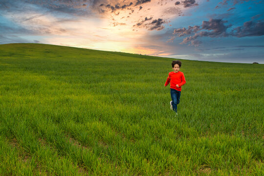 6 Year Old Boy Running Through A Green Meadow With A Spectacular Sky And A Nice Horizon. Image With Copy Space. Freedom Concept