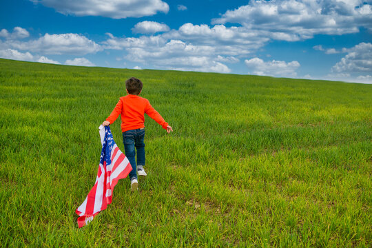 Child Running And Enjoying Himself In A Green Meadow With A Dramatic Sky. He Bears The Flag Of The United States, Wears A Red T-shirt And Jeans. Image With Copy Space. Nature And People.