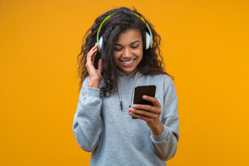 Portrait of cute smiling student girl wearing wireless headphones and browsing tracks on her smartphone, isolated over yellow background