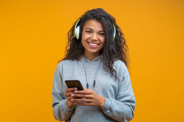 Portrait of casually dressed beautiful girl with charming smile wearing wireless headphones and posing over yellow background with mobile phone in hands - obrazy, fototapety, plakaty
