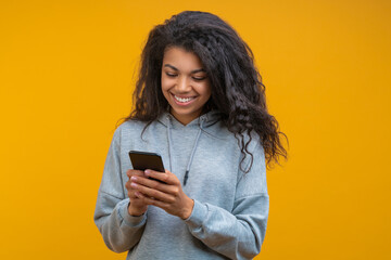 Half-length studio portrait of beautiful casually dressed dark skinned girl with perfect charming smile texting message via her mobile phone isolated on bright colored yellow background