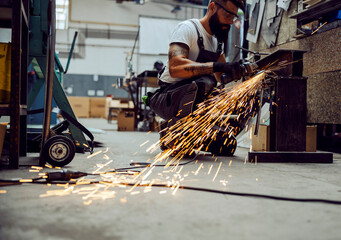 A worker is crouching in the workshop and grinding metal parts. 