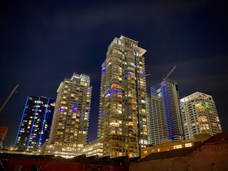 Group of high-rise towers of apartments in Downtown Los Angeles, some under construction, lit up with different colors at night