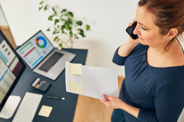 Woman entrepreneur having business  conversation on mobile phone. Businesswoman working in office holding documents and smartphone. Woman standing by desk with computers