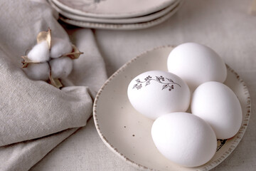 White Easter eggs on beige plates, with a minimal black sprig pattern. There is a linen tablecloth and a cotton napkin on the table. Natural materials, minimal concept. Place for text.