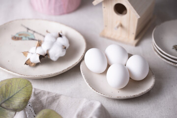 Fototapeta na wymiar White Easter eggs on beige plates near the birdhouse and with cotton on a saucer on the table. Natural materials, minimal concept. Place for text.