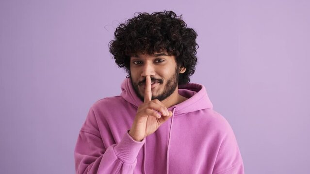 Happy curly-haired Indian man looking around and showing shh gesture in the purple studio