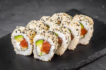 Foto op Plexiglas appetizing california sushi roll with cheese avocado cucumber and salmon in sesame seeds on a black stone plate © Narsil