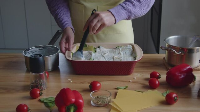 Woman is cooking tasty dinner at home, spreading white sauce on lasagna noodles, preparing meal to bake in own, Slow motion.