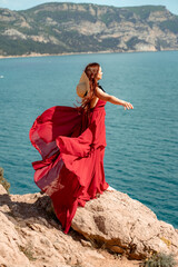 A woman, a side view in a red flying dress fluttering in the wind, a girl in a fluttering dress on the background of the sea. A straw hat hangs at the back of the neck. The concept of a vacation at se