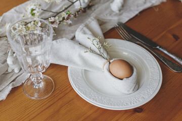 Easter egg in bunny napkin on plate with cutlery, bunny, spring flowers and rustic cloth on wooden table. Stylish Easter brunch table setting. Holiday arrangement and eco friendly decorations