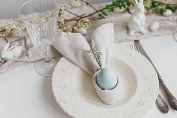 Happy Easter! Stylish elegant Easter brunch table setting. Easter egg in bunny napkin on plate with cutlery, bunny, spring flowers on rustic white table. Modern eco table arrangement