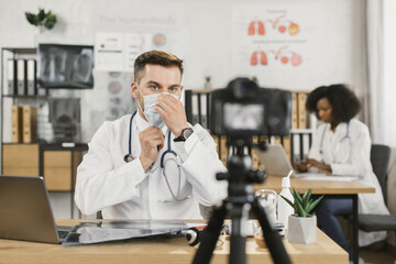 Experienced therapist sitting at hospital cabinet and recording video on camera while explaining about protection from face mask wearing. Concept of pandemic, healthcare and technology.