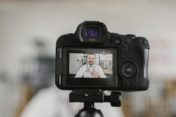 View through camera on positive caucasian doctor in white lab coat sitting at desk with various pills in hands. Competent therapist recording video about health treatment.