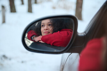 Reflection in the car side view mirror of a beautiful confident serene European woman, sitting on a driver seat and admiring beautiful snowy woodland while travelling by car