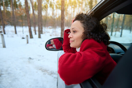 Side portrait. Curly dark-haired serene woman relaxing sitting on a driver seat of her car, resting in the snow covered woodland with beautiful sunbeams falling into the forest path. Winter travel