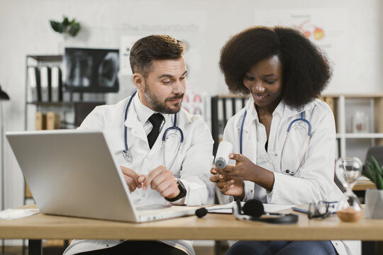 Multiracial male and female doctors using modern laptop for online seminar. Two medical workers sitting at desk and talking about usage of contactless infrared thermometer.