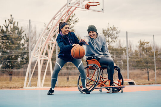 Happy woman and her friend in wheelchair plays basketball on outdoor sports court.