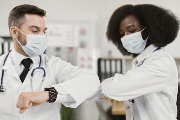 Two multiracial male and female doctors in face masks doing elbow bump during meeting a hospital...