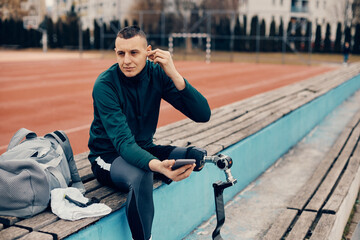 Athletic man uses smart phone and wireless earphones while relaxing at the stadium after the practice.
