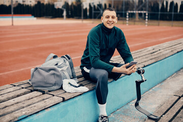 Happy athlete with a prosthetic leg uses smart phone and wireless earphones while relaxing at the stadium