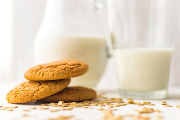 Pile oatmeal cookies and oat cereal flakes on white table on milk glass jug background