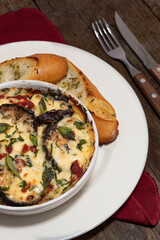 Lasaña lista para comer de berenjena, queso mozzarella, queso parmesano, albahaca, perejil y pan de ajo en plato blanco sobre un fondo de madera  y junto a unos cubiertos