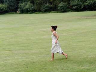 Portrait of young Asian woman running on grass field with barefoot in forest park, beautiful Chinese girl in white dress enjoy her carefree time in sunny summer day, full length shot.