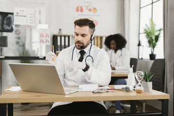 Qualified doctor in lab coat and gloves holding sterile syringe and talking during video conference on laptop. Male therapist in headset having online appointment with patient.