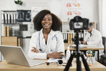 Smiling african american woman in lab coat sitting at desk with modern laptop and using camera for recording video blog. Qualified female doctor creating content for her medical channel.