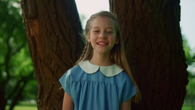 Joyful little girl pose at tree trunk. Smiling kid with braid look in camera.