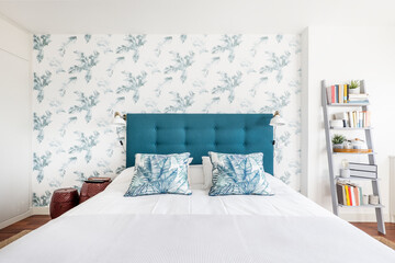 Headboard upholstered in blue capitone fabric with matching cushions with floral motifs, decorative paper on the wall and a shelf with a book in one corner