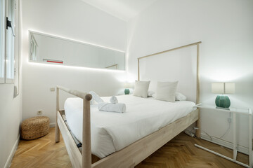 Bedroom with wooden bed with railings, white quilt, large long mirror and lamps on the bedside tables