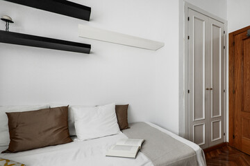 Open book on small bed in bedroom with brown and white cushions, gray courtesy blanket and white built-in wardrobe