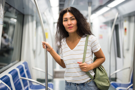 Asian woman with shoulder bag standing in subway train, holding handrail and looking in camera.