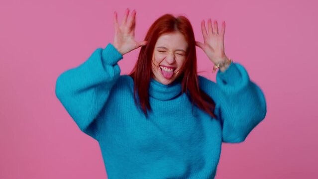 Funny comic teen student girl 20s years old in blue sweater making playful silly facial expressions and grimacing, fooling around showing tongue. Young adult woman isolated on pink wall background