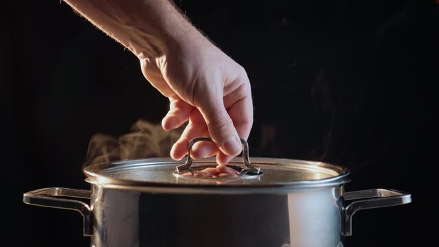 Male Caucasian hand putting on a cover on the steaming stainless steel pot with boiling water, isolated on a black background.