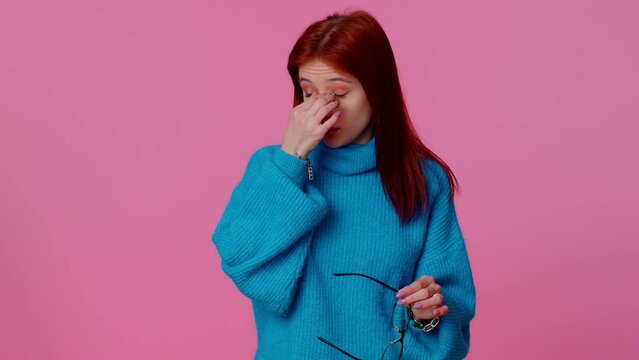 Portrait of exhausted tired redhead woman takes off glasses, feels eyes pain, being overwork burnout from long hours working, wearing blue sweater. Indoors studio shot isolated on pink background