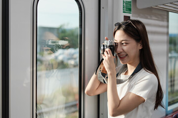 A Beautiful Asian female tourist with camera in the passenger cabin, traveling by sky train, taking snapshot photos when transporting in urban view, city lifestyle by railway, happy journey vacation.