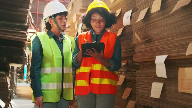 Two female workers and colleagues in safety uniforms and helmet stock check, control production in storage of warehouse factory with a lot of paper stack, friend works in recycle manufacture business.