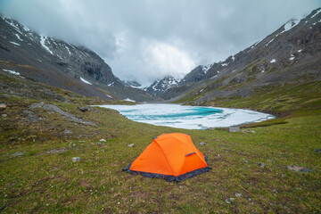 Atmospheric mountain landscape with orange tent near frozen alpine lake and snowy mountains. Awesome scenery with icy mountain lake on background of snow mountains in low clouds. Tent near ice lake.