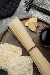 Fototapeta na wymiar Different uncooked rice noodles, soy sauce, cut ginger and chopsticks on grey table, flat lay